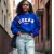 Femme souriante, portant le sweat EmakoBollo en couleur Team Royal, imprimé EmakoBollo, posant dans un cadre urbain décontracté