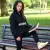 Jeune fille, portant le sweat à capuche noir imprimé "EmakoBollo" en noir, assise confortablement sur un banc de parc, avec un livre à la main.