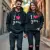 Un couple jeune, un homme et une femme, souriant et se tenant la main, portant chacun le sweat à capuche unisexe "I love you" noir, marchant dans une rue urbaine ensoleillée. Style streetwear, photo de haute qualité.