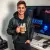 Un jeune homme (geek) souriant, tenant le mug café "Game Over" près de son écran d'ordinateur, une tasse fumante à la main.