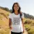 Une jeune femme souriante, portant le T-shirt de sport blanc, en pleine randonnée sur un sentier ensoleillé. Le t-shirt a l'air léger et respirant, et elle semble à l'aise et pleine d'énergie.