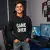 un jeune homme souriant, portant le pull à capuche noir "Game Over", assis devant un setup de gaming stylé